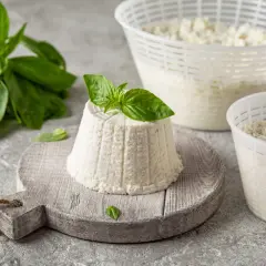 Świeży serek ricotta na desce, ozdobiony bazylią. Obok dwa pojemniki z serkiem i gałązka bazylii.