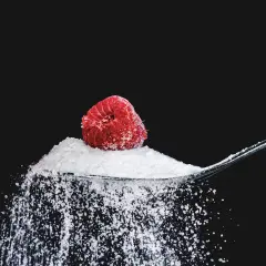 Malina na łyżce z erytrytolem, który sypie się na czarne tło. Erytrytol to naturalny słodzik.