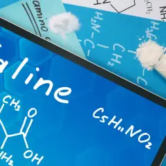 Tablet z napisem "Valine" i wzorem chemicznym. Obok probówka z białym proszkiem, symbolizującym tę aminokwas.