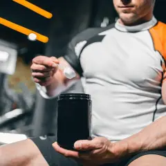 Mężczyzna w sportowej koszulce trzyma czarny pojemnik i miarkę z białym proszkiem. To jego codzienny stack kreatynowy.