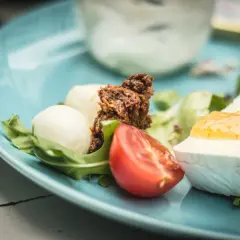 Pyszne śniadanie keto: ser camembert z konfiturą, jajka przepiórcze, pomidor i rukola na błękitnym talerzu.