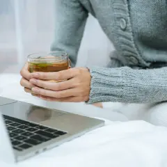 Kobieta w szarym swetrze pije herbatę, zastanawiając się, l teanina co to. Obok leży laptop.