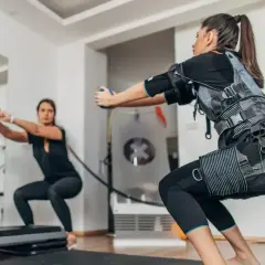 Kobiety w kamizelkach EMS wykonują przysiady, ćwicząc budowę mięśni.