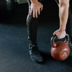 Mężczyzna w czarnych legginsach i butach sportowych podnosi kettlebell. Jego nogi są napięte, gotowe do treningu.