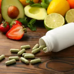 Kapsułki multiwitaminy rozsypane obok białej buteleczki, na tle świeżych owoców: kiwi, awokado, truskawek, cytrusów i granatu.