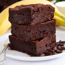Stos puszystych brownie z banana i kakao, ozdobiony ziarnami kawy, na talerzyku z bananem w tle.