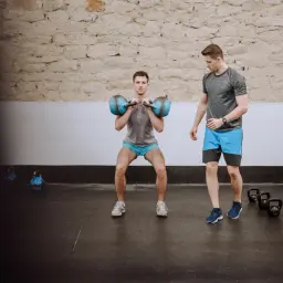 Trener obserwuje mężczyznę wykonującego przysiady z dwoma kettlebellami, ćwicząc na **hipertrofię** mięśni.