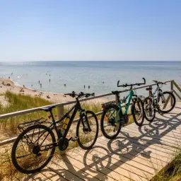 Rowerowa trasa nad morzem. Kilka rowerów czeka na drewnianej kładce przy plaży, gdzie widać kąpiących się ludzi.