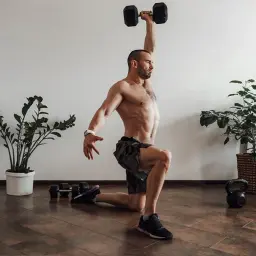 Mężczyzna wykonuje ćwiczenie z hantlem nad głową, element treningu crossfit. Obok leżą hantle i kettlebell.