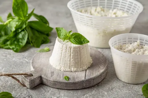 Świeży serek ricotta na desce, ozdobiony bazylią. Obok dwa pojemniki z serkiem i gałązka bazylii.