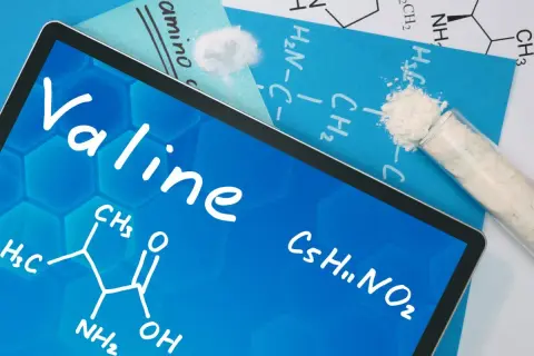 Tablet z napisem "Valine" i wzorem chemicznym. Obok probówka z białym proszkiem, symbolizującym tę aminokwas.