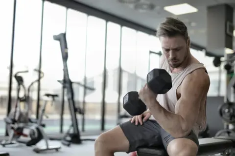 Mężczyzna ćwiczy biceps z hantlem, doskonaląc swoją biomechanikę. W tle siłownia z maszynami.