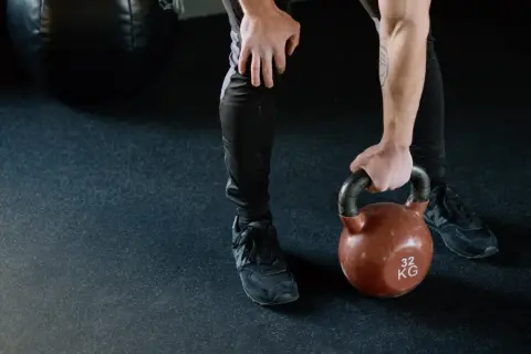 Mężczyzna w czarnych legginsach i butach sportowych podnosi kettlebell. Jego nogi są napięte, gotowe do treningu.
