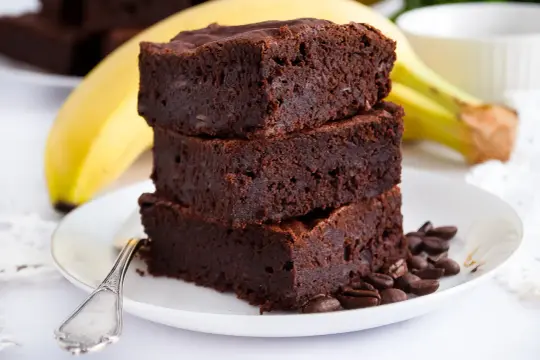 Stos puszystych brownie z banana i kakao, ozdobiony ziarnami kawy, na talerzyku z bananem w tle.
