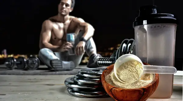 Mięśniak odpoczywa po treningu z odżywką carbo w miseczce i shakerem. Obok leżą hantle i talerze.