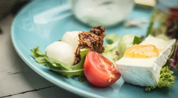 Pyszne śniadanie keto: ser camembert z konfiturą, jajka przepiórcze, pomidor i rukola na błękitnym talerzu.