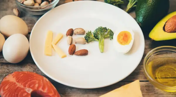 Czy dieta keto jest zdrowa? Na talerzu litery KETO z sera, orzechów, brokuła i jajka. Wokół łosoś, awokado, oliwa.