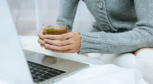 Kobieta w szarym swetrze pije herbatę, zastanawiając się, l teanina co to. Obok leży laptop.