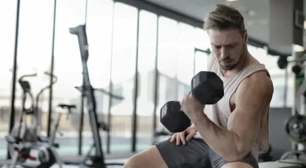 Mężczyzna ćwiczy biceps z hantlem, doskonaląc swoją biomechanikę. W tle siłownia z maszynami.