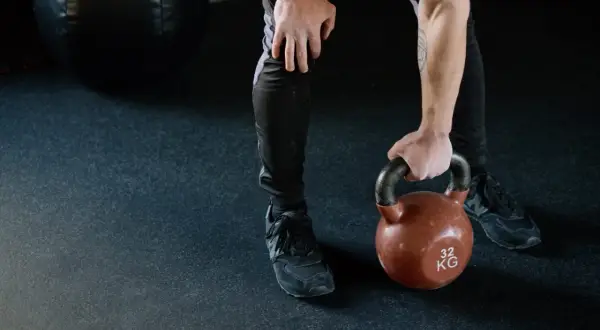Mężczyzna w czarnych legginsach i butach sportowych podnosi kettlebell. Jego nogi są napięte, gotowe do treningu.