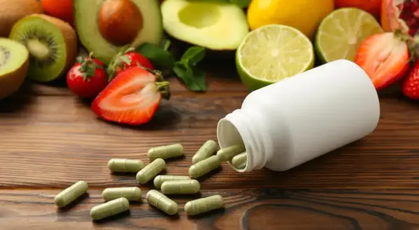 Kapsułki multiwitaminy rozsypane obok białej buteleczki, na tle świeżych owoców: kiwi, awokado, truskawek, cytrusów i granatu.