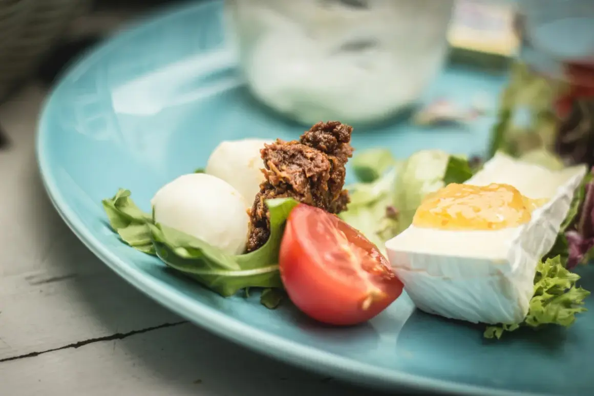 Pyszne śniadanie keto: ser camembert z konfiturą, jajka przepiórcze, pomidor i rukola na błękitnym talerzu.