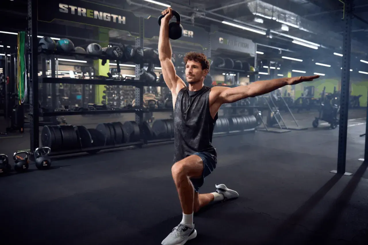 Mężczyzna wykonuje wykrok z kettlebell nad głową, ćwicząc siłę i stabilność. W tle widoczne są półki ze sprzętem treningowym.