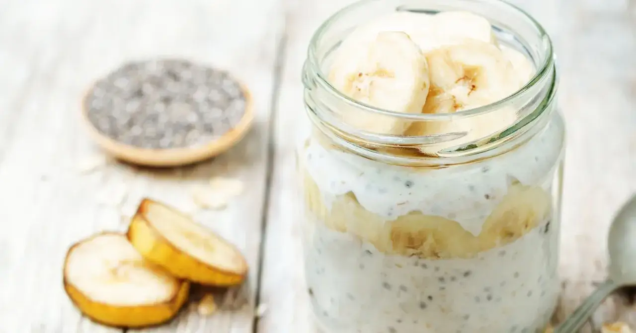 Słoik z warstwową nocną owsianką z bananem, nasionami chia i płatkami owsianymi. Obok leżą plasterki banana.