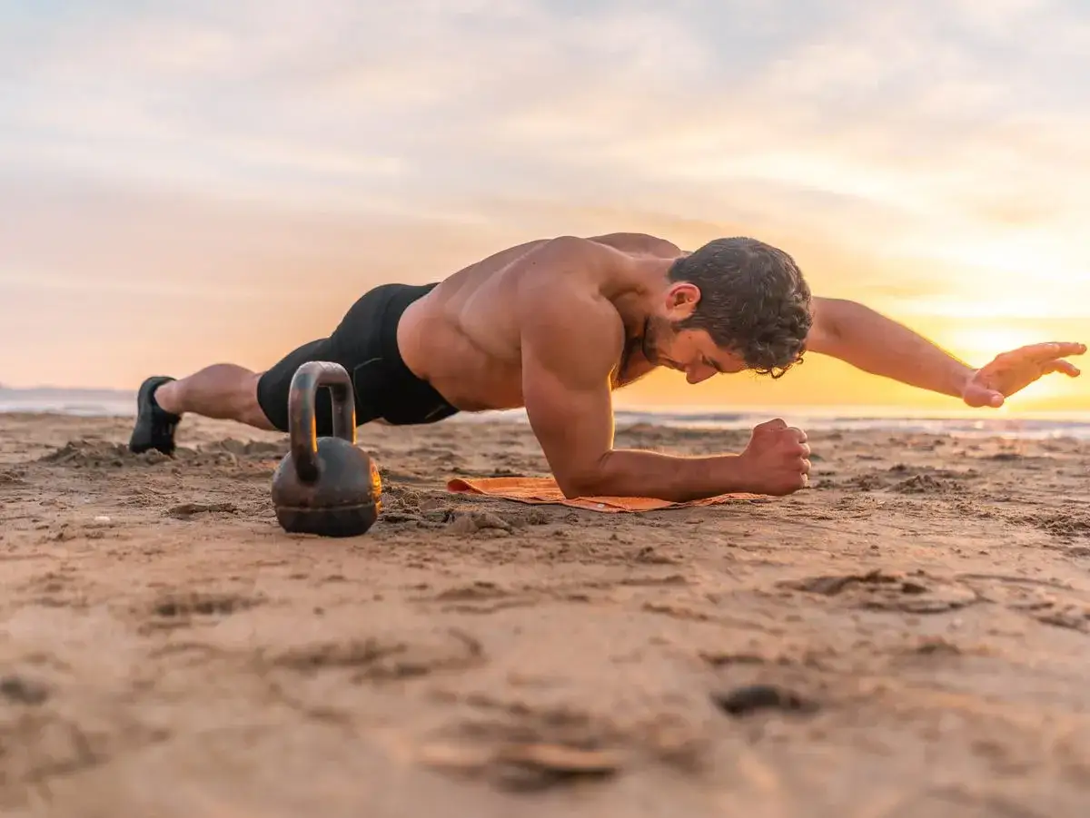 Mężczyzna wykonuje ćwiczenia na mięśnie głębokie brzucha na plaży. Kettlebell obok. Zachód słońca.