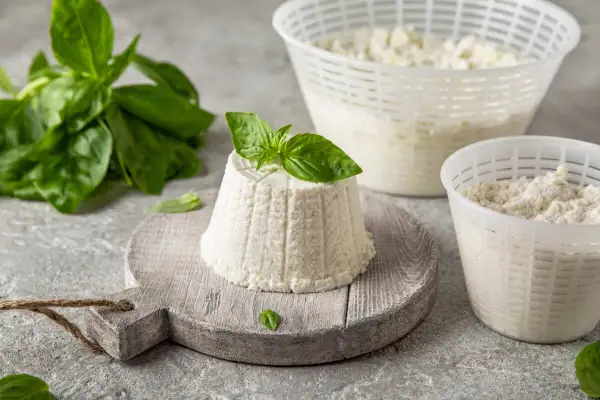 Świeży serek ricotta na desce, ozdobiony bazylią. Obok dwa pojemniki z serkiem i gałązka bazylii.