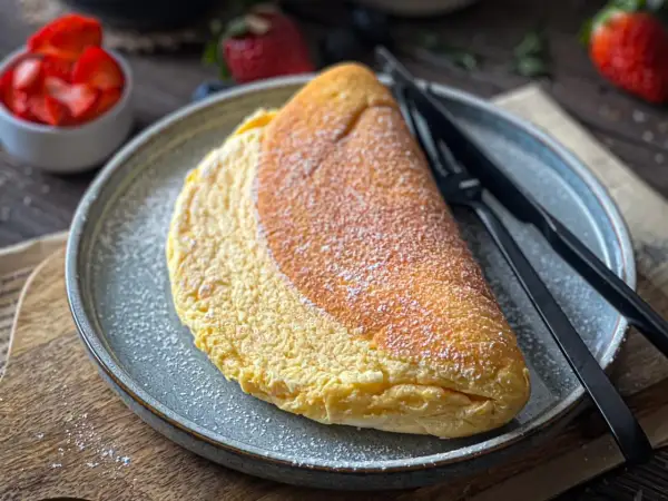 Puszysty omlet z białek, lekko przyprószony cukrem pudrem, czeka na talerzu obok świeżych truskawek.