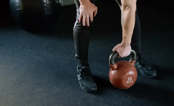 Mężczyzna w czarnych legginsach i butach sportowych podnosi kettlebell. Jego nogi są napięte, gotowe do treningu.