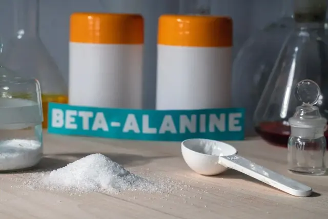 Biały proszek, miarka i etykieta z napisem BETA-ALANINE na tle laboratoryjnego sprzętu.
