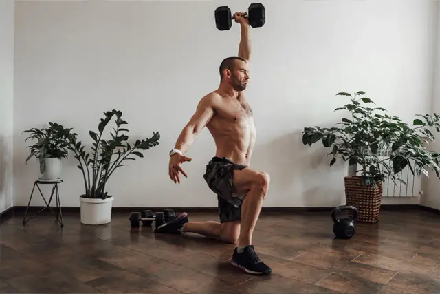 Mężczyzna wykonuje ćwiczenie z hantlem nad głową, element treningu crossfit. Obok leżą hantle i kettlebell.